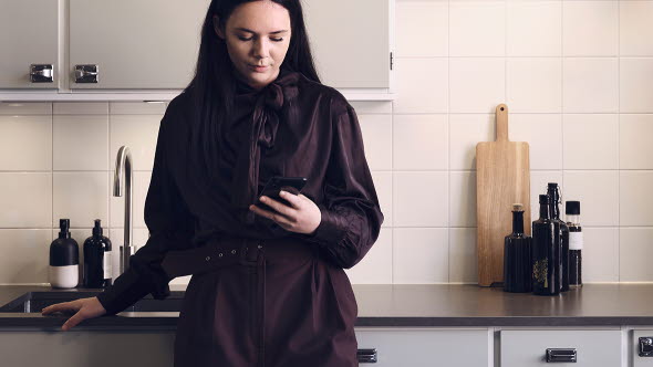 Kvinna som står i sitt kök och tittar på sin mobil